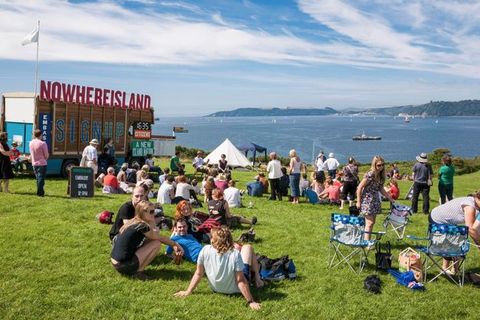 People gather on a grassy seaside near a stand with signs reading, “Nowhereisland”, “Sign up”, and “Embassy”.