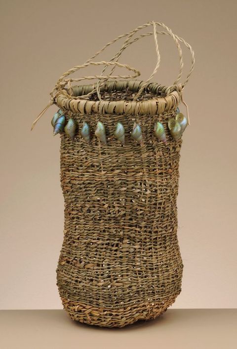 Basket. Tall woven vessel has a rounded bottom and iridescent, spiralling shells around the mouth. Thin handles loop up from the top.