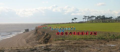 Installation. Steep, low cliffs face a beach to our left. Coloured flags are planted in lines moving back from the edge of the cliff. Each flag has a letter on it. The letters spell out words, and the flags that make up a word have the same colour. Together the flags read, “Submission is advancing at a frightful speed”.